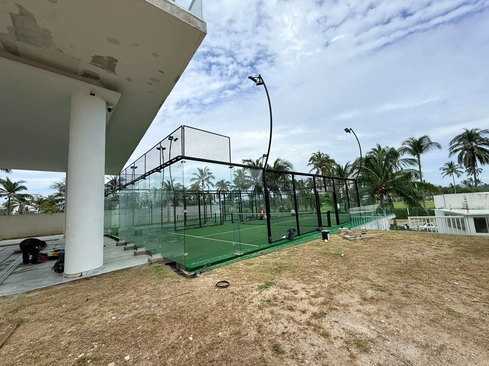 Vista general de una cancha de pádel con cinturón perimetral rodeada de palmeras junto a un edificio