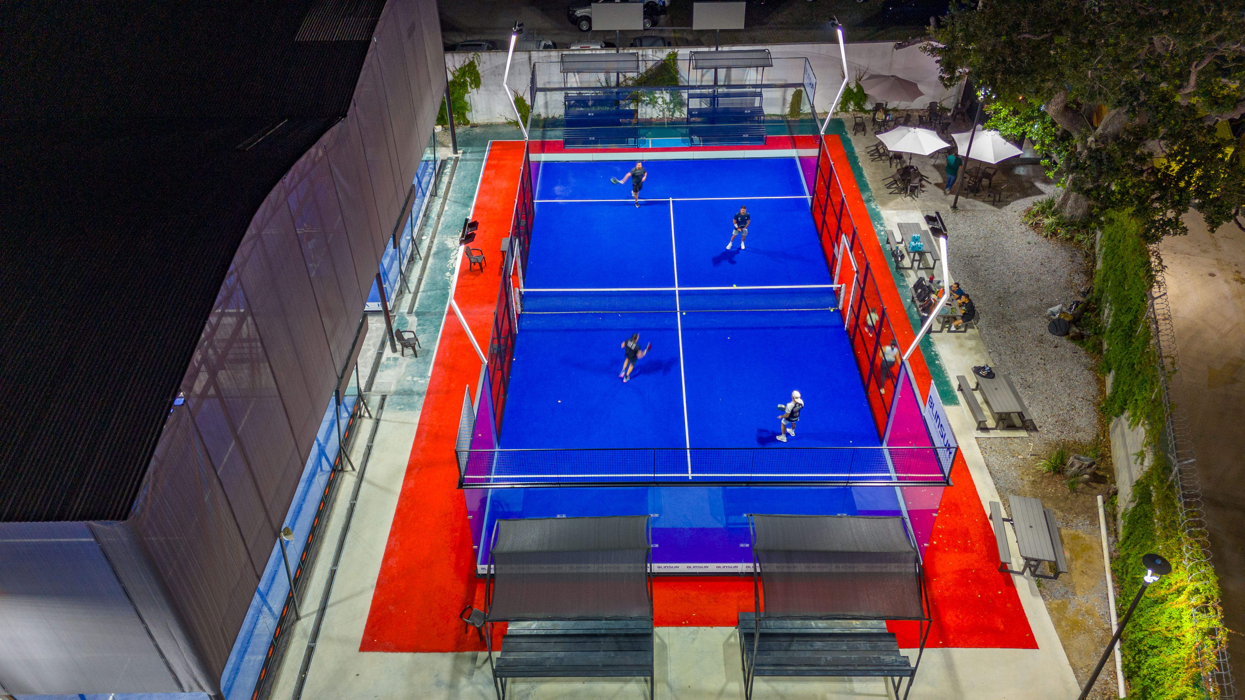 Cancha de pádel azul con pasillos rojos vista desde dron durante partido nocturno