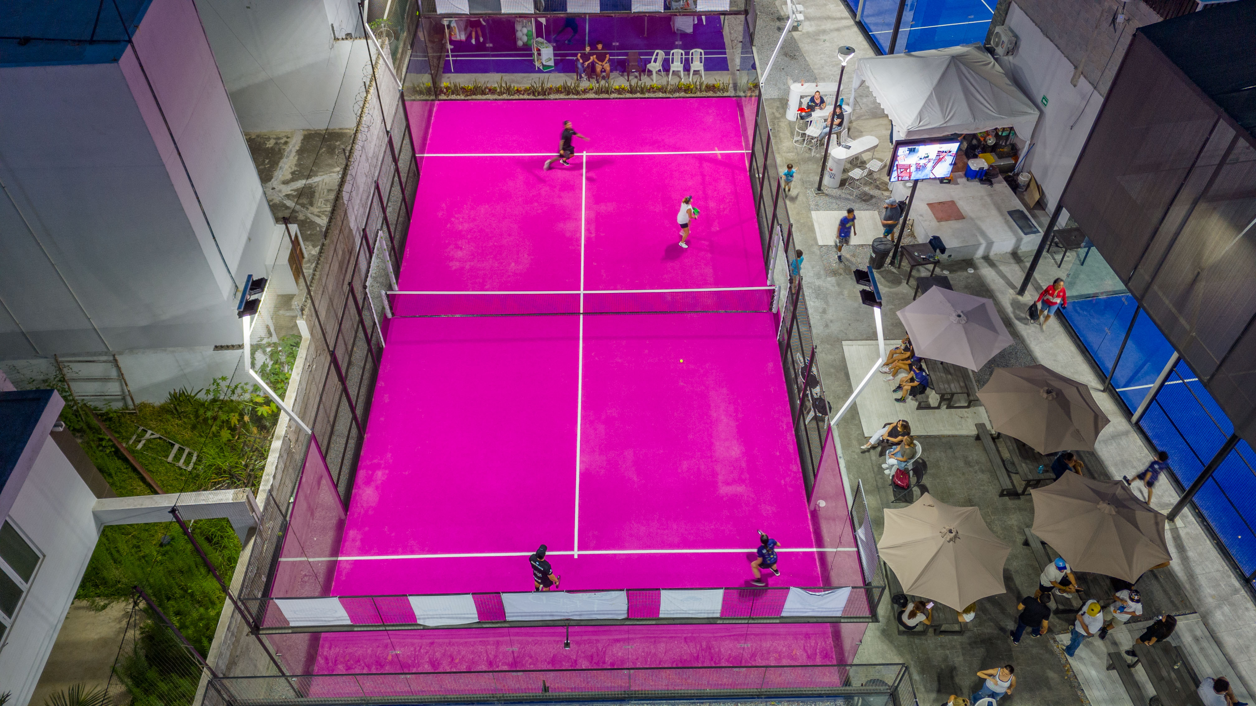 Cancha de pádel rosa vista aérea con jugadores y zona social